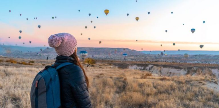 A woman wearing a cozy hat and backpack, looking at multiple hot air balloons in the air