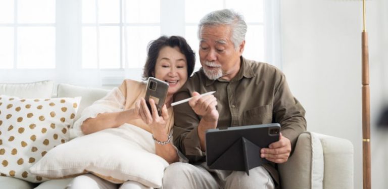 An older couple sitting on a couch together. One is holding a phone, the other is holding a tablet.