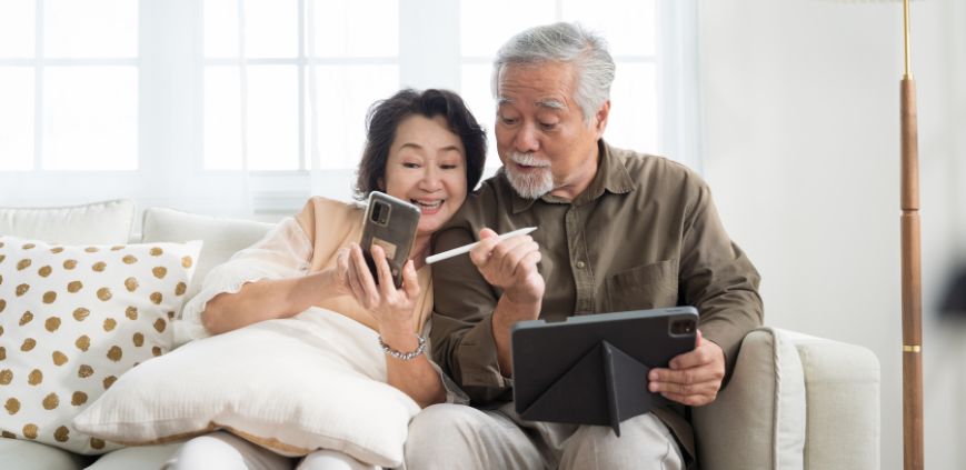 An older couple sitting on a couch together. One is holding a phone, the other is holding a tablet.